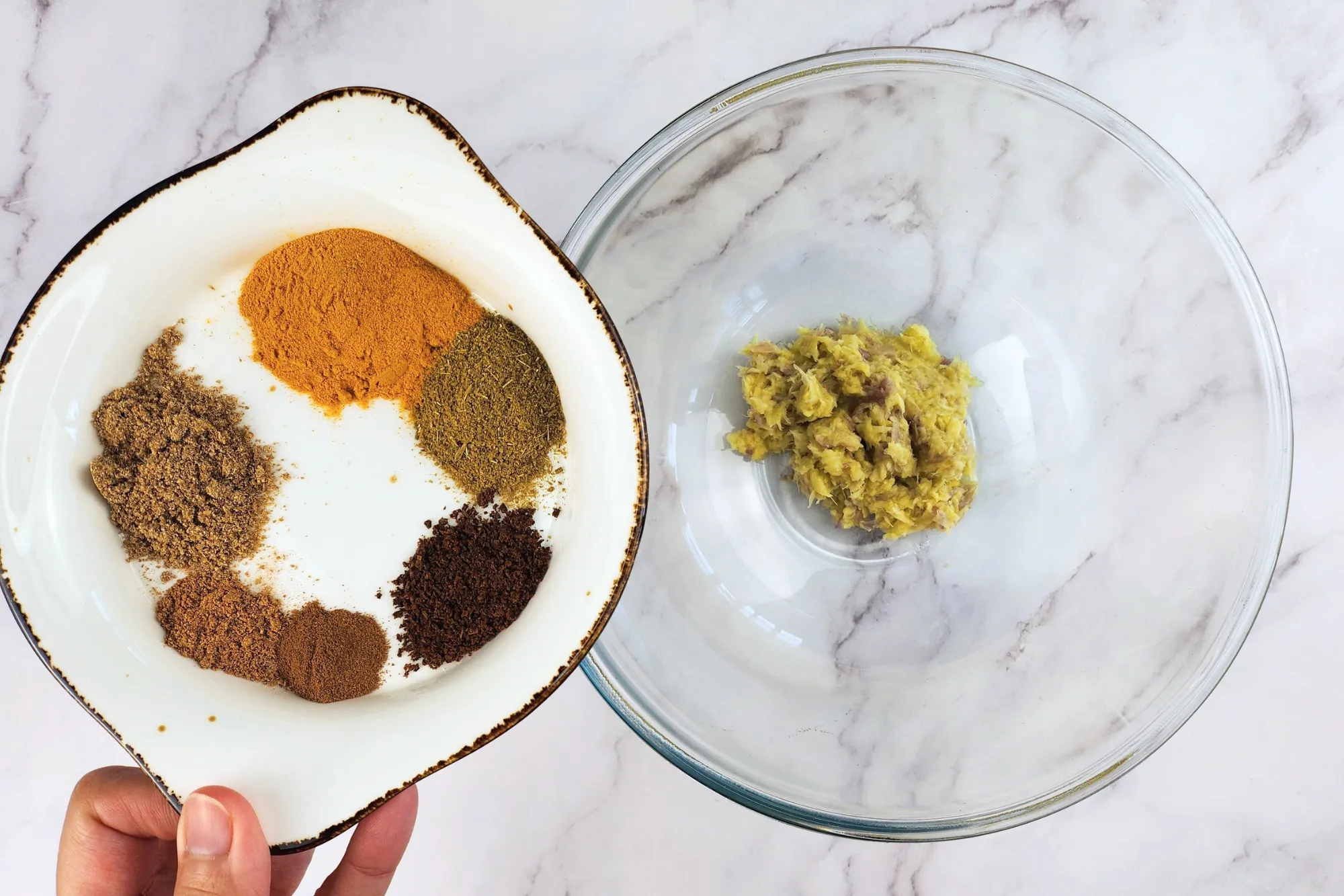 A hand is holding a plate of spices above a mixing bowl with minced herbs.