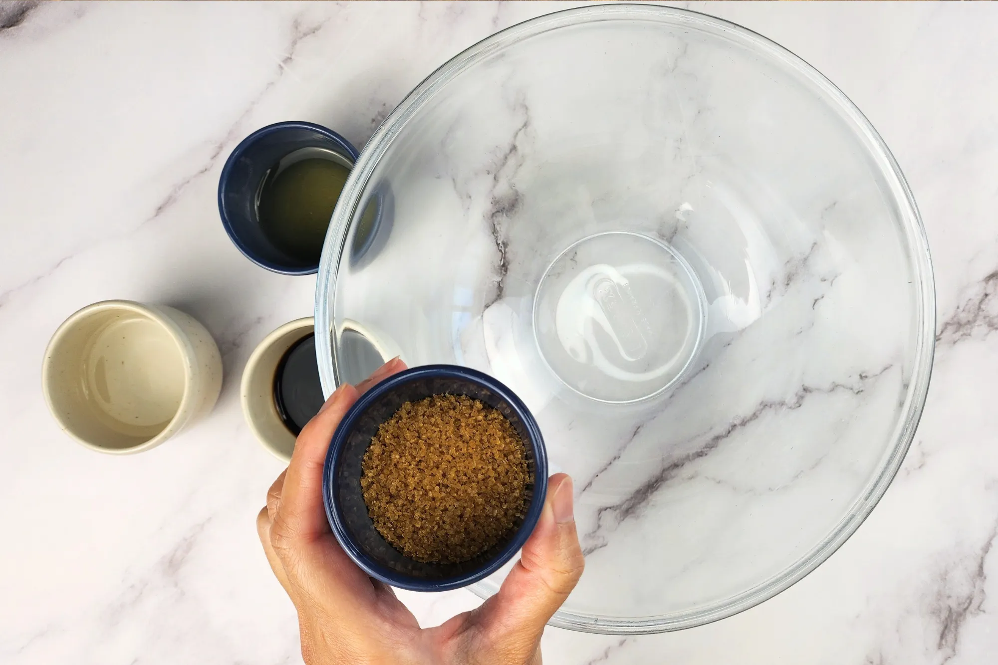 A hand is holding a small bowl of brown sugar above a mixing bowl.