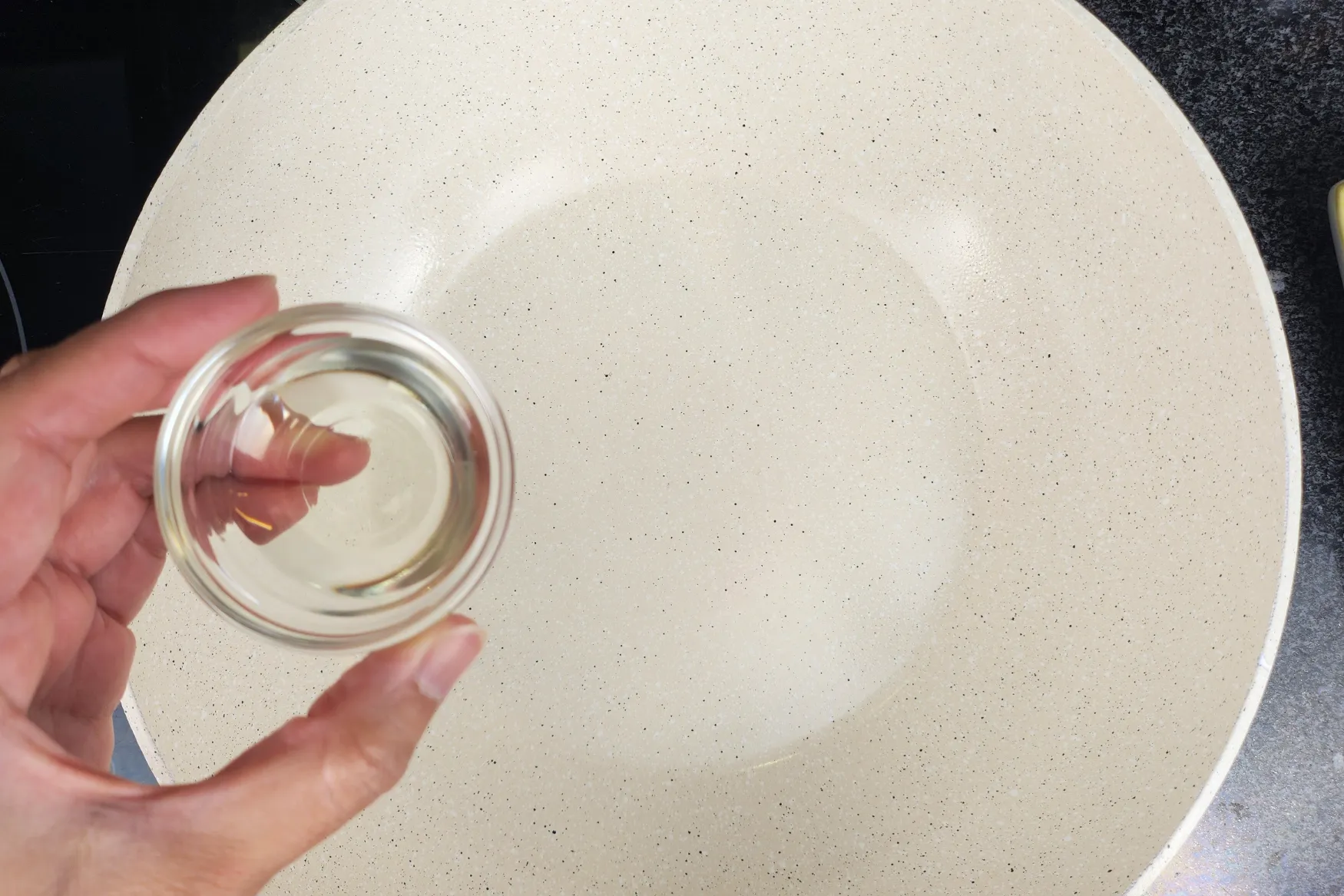 A hand is holding a small bowl of cooking oil above a wok