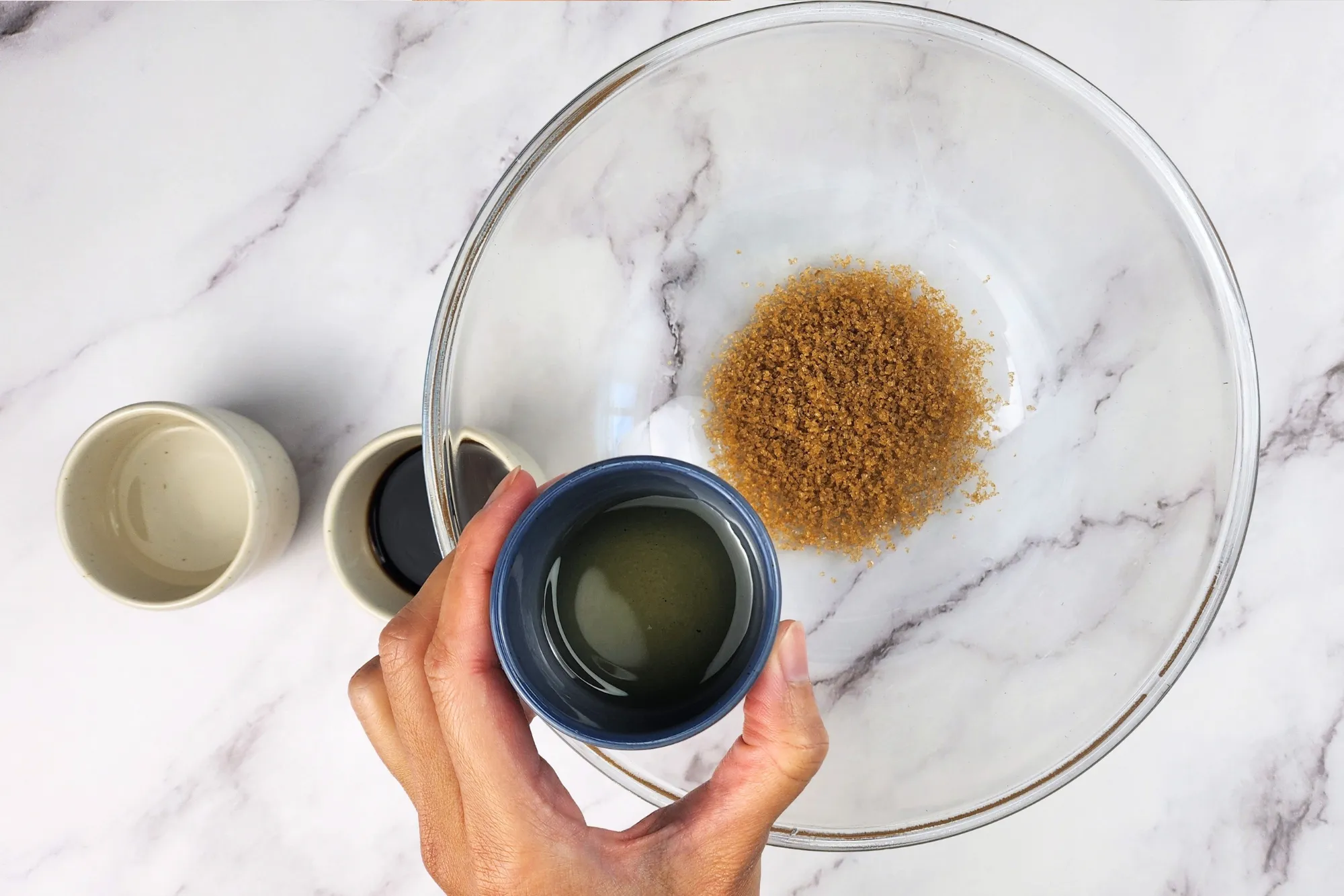 A hand is holding a small bowl of mirin above a mixing bowl.
