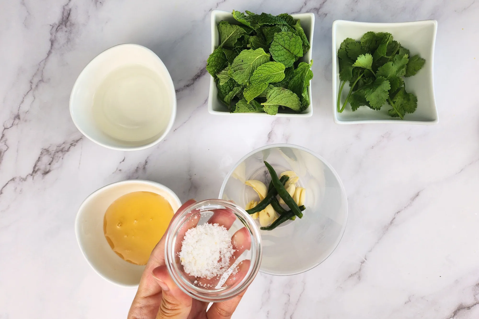 A hand is holding a small bowl of salt is above a mixing jar surrounded with mint, cilantro, vinegar and honey.