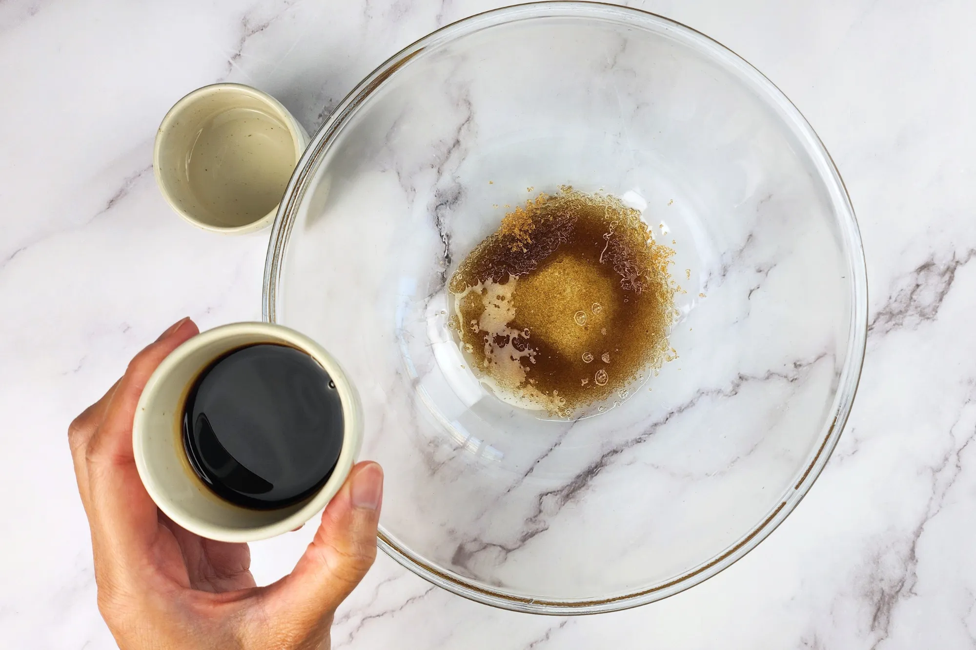 A hand is holding a small bowl of soy sauce above a mixing bowl.