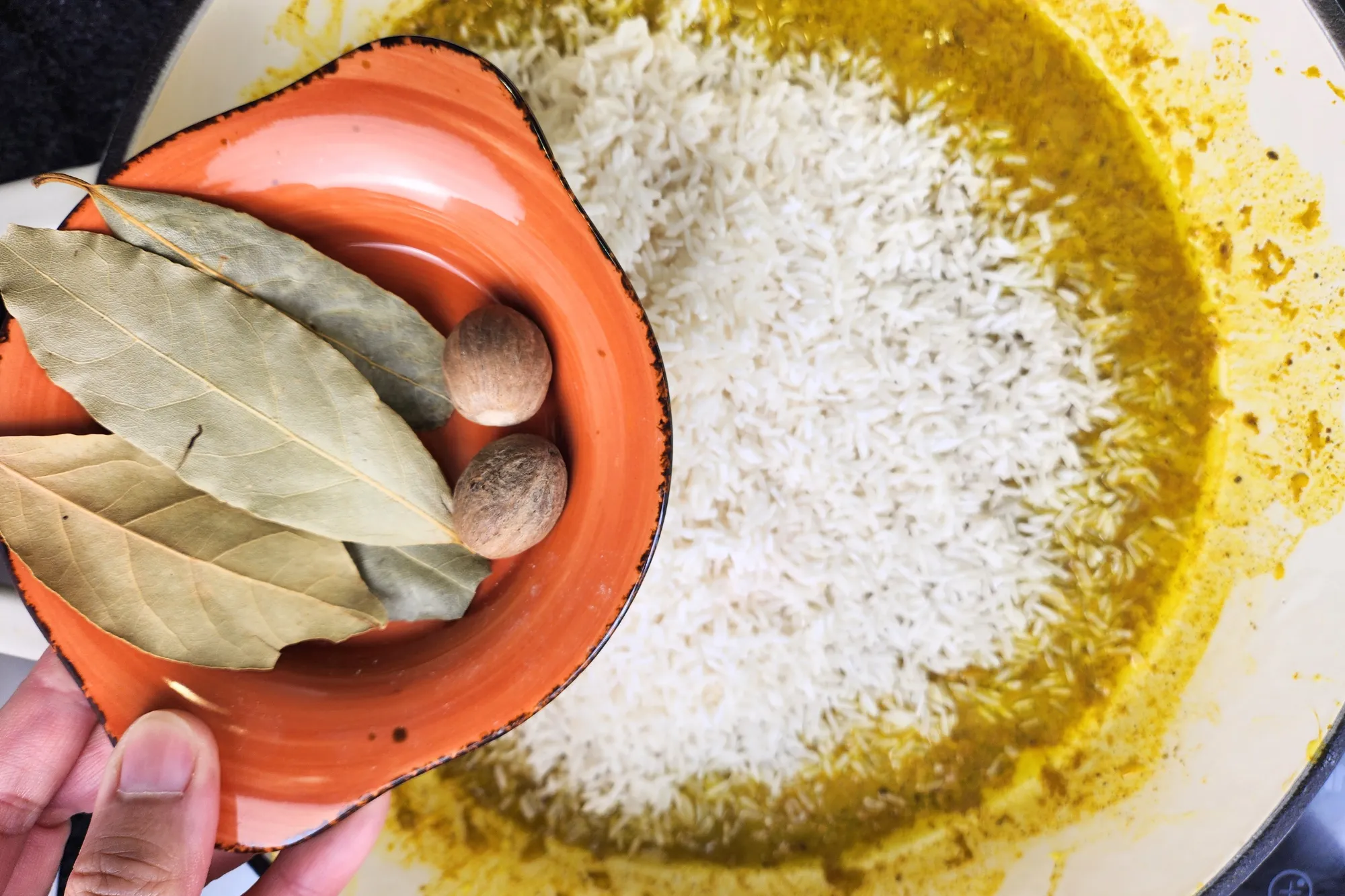 A hand is holding a small plate with bay leaves and Cardamom Pods above a pot with uncooked rice with yellow sauce.