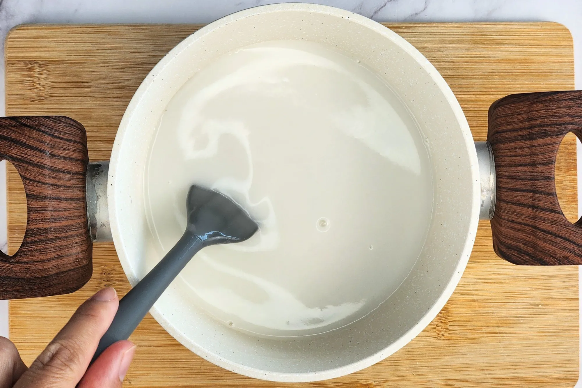 A hand is holding a spatula in a pot of white liquid on the wooden board