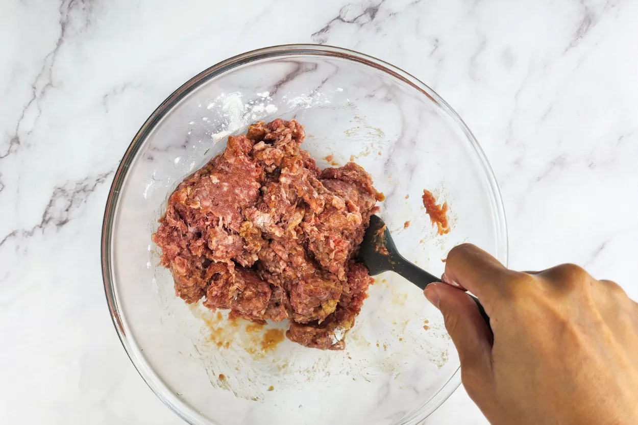 A hand is holding a spatula mixed ground beef in a mixing bowl.