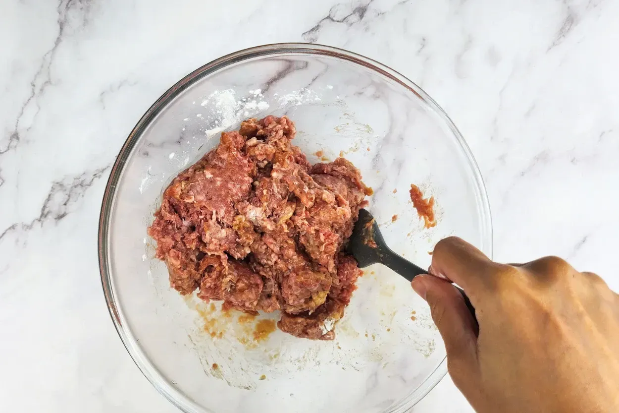 A hand is holding a spatula mixed ground beef in a mixing bowl.