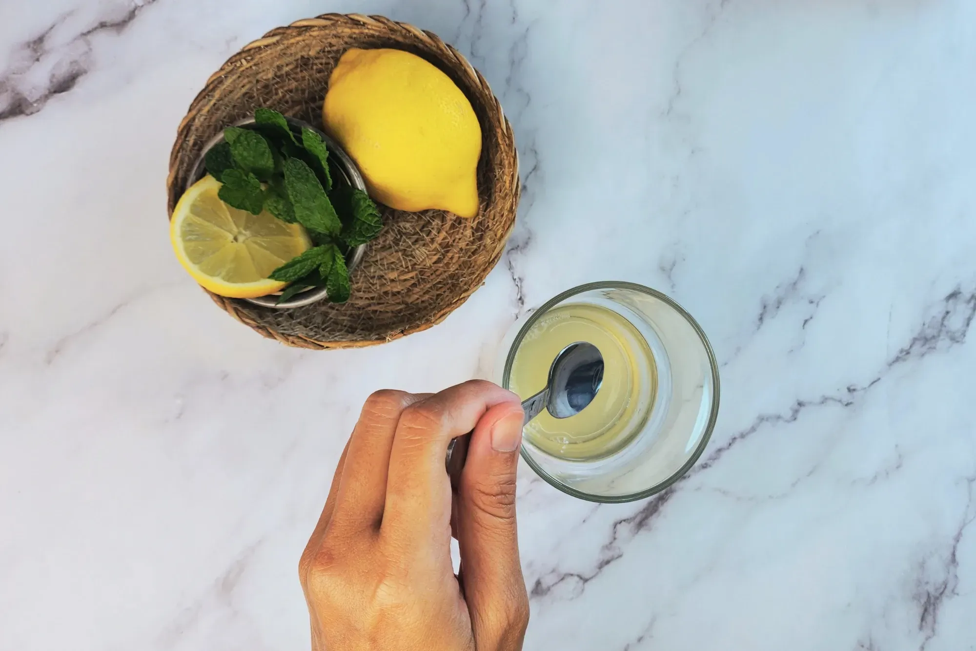 A hand is holding a spoon above a glass next to lemon and mint.