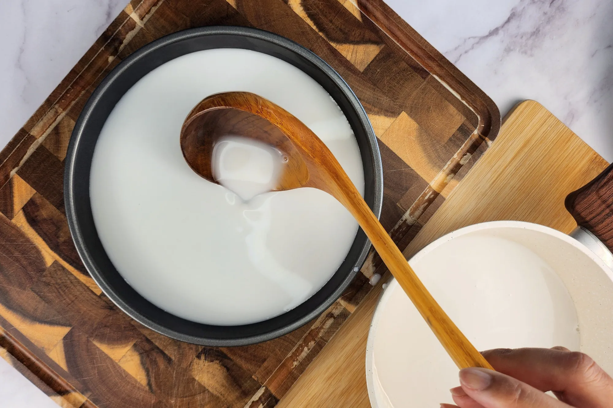 A hand is holding a wooden spoon pouring white liquid to the tray on the chopping board