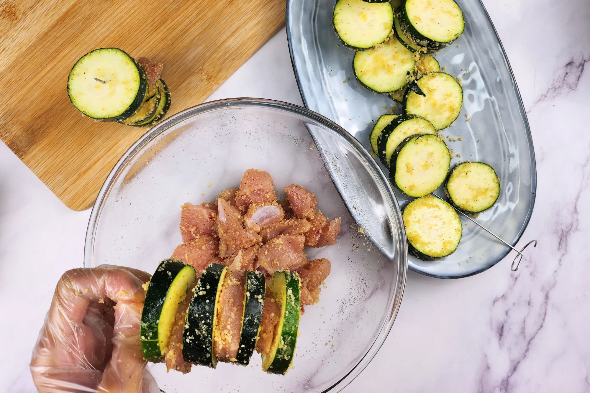 A hand is holding a zucchini skewers above a mixing bowl with chicken and zucchinis on a plate.