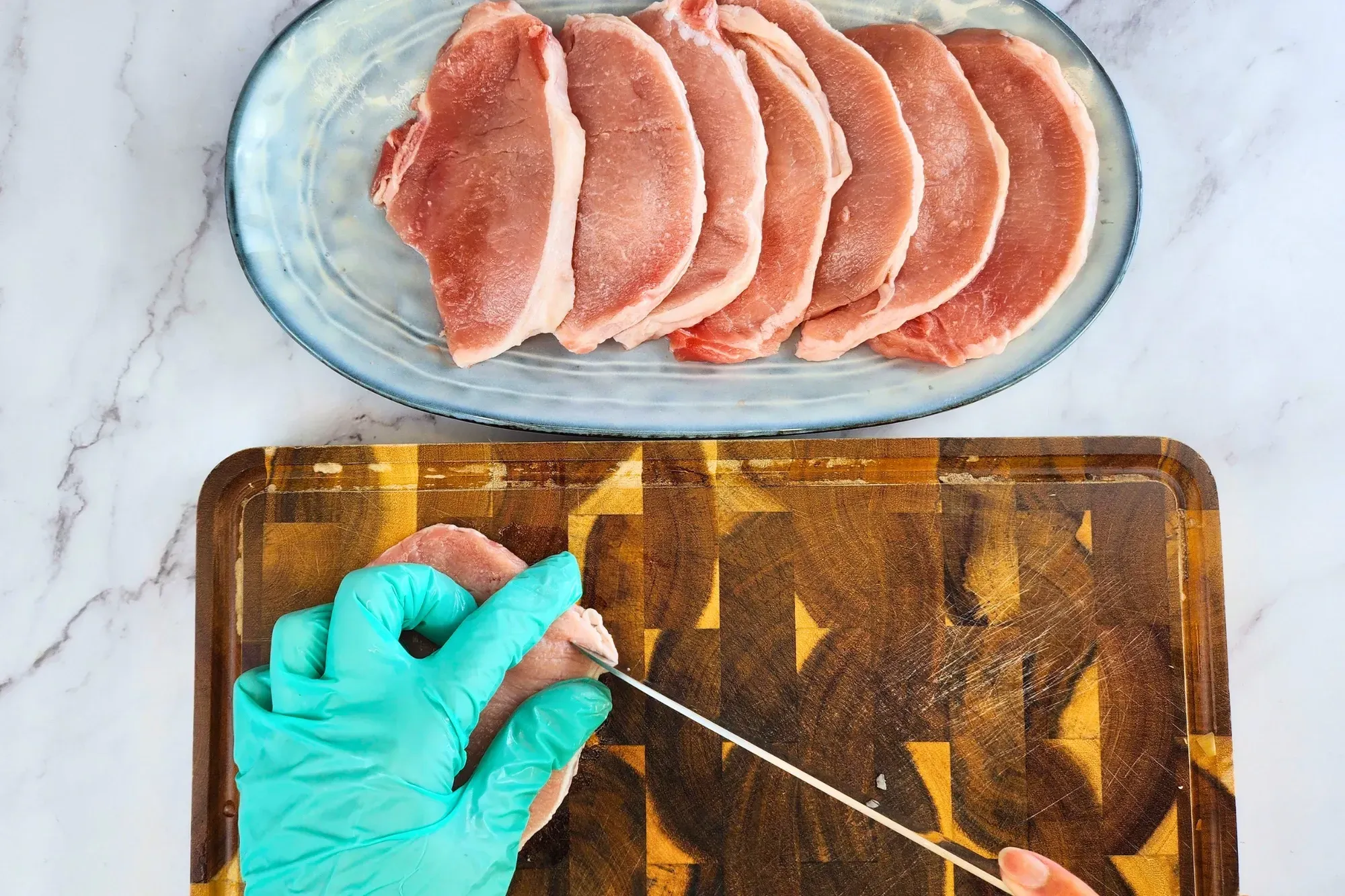 A left hand hold a pork chop with a right hand cut a pork chop with a plate of raw pork chops