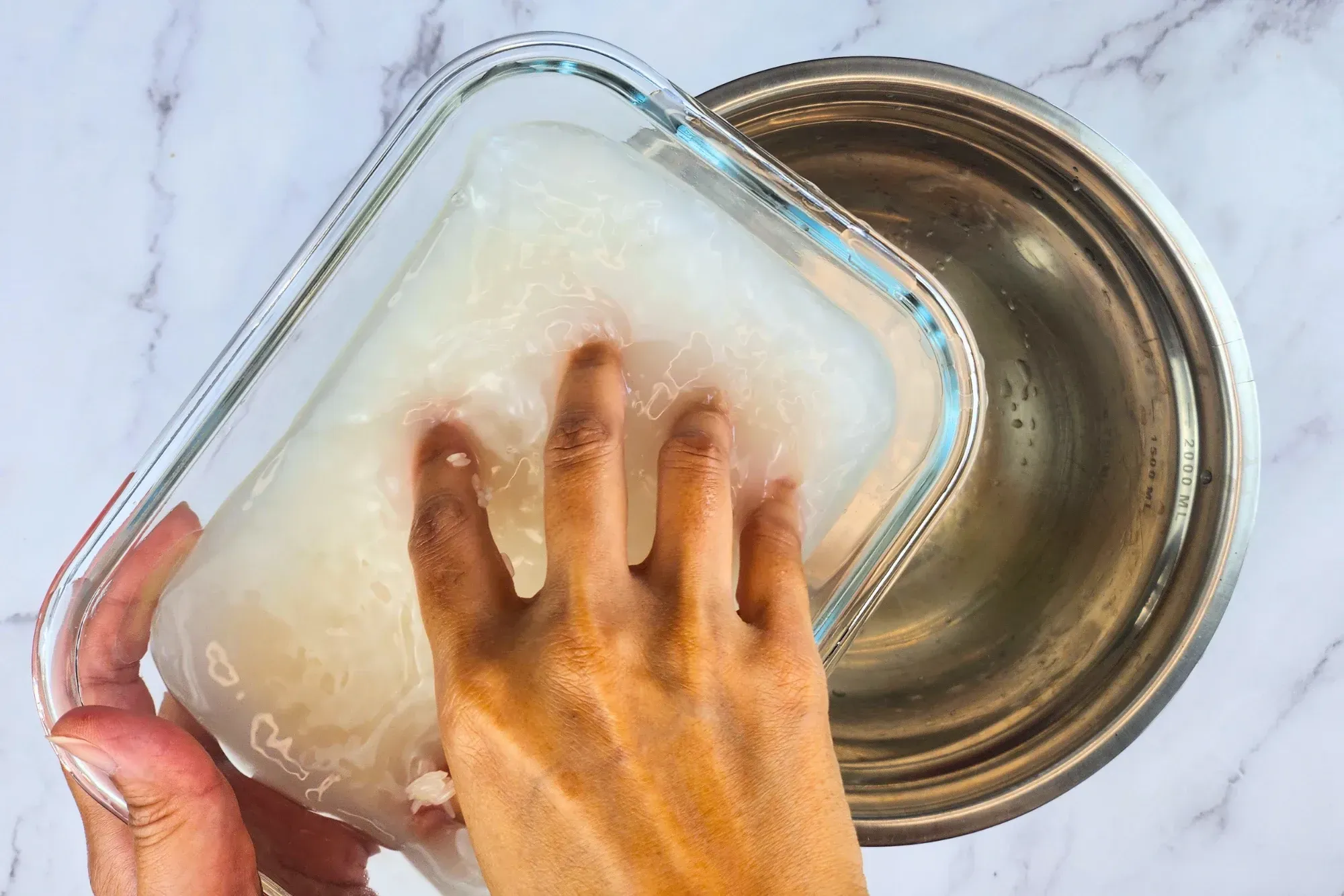 A left hand is holding a container of jasmine rice with water and a right hand is rubbing jasmine rice above a bowl