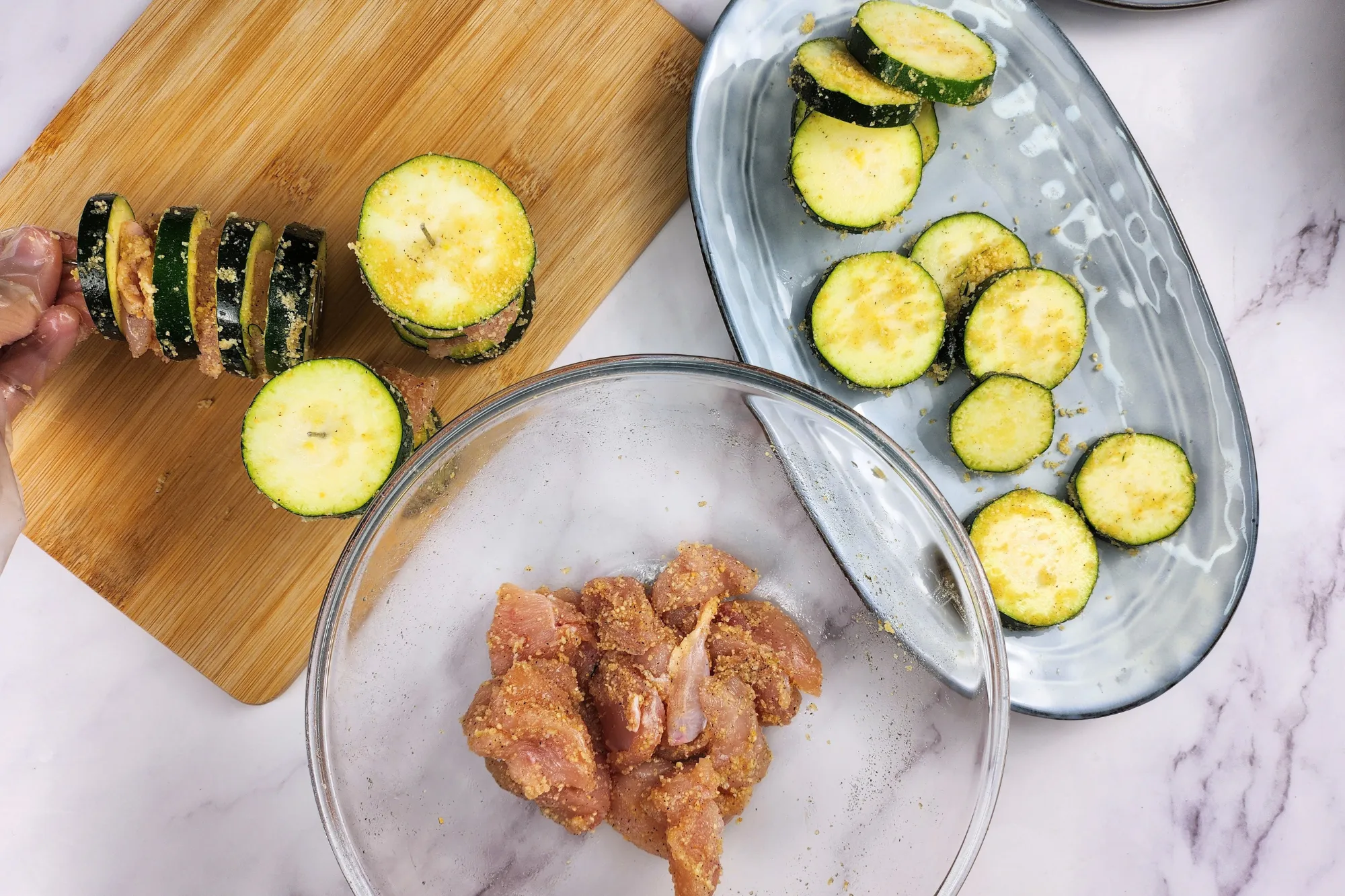 A mixing bowl with chicken and zucchinis on a plate and fews of zucchini skewers on a wooden board.