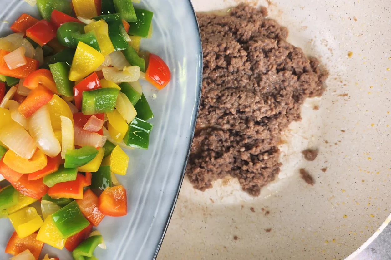 A plate of cooked bell peppers and onions above a wok with cooked ground beef.