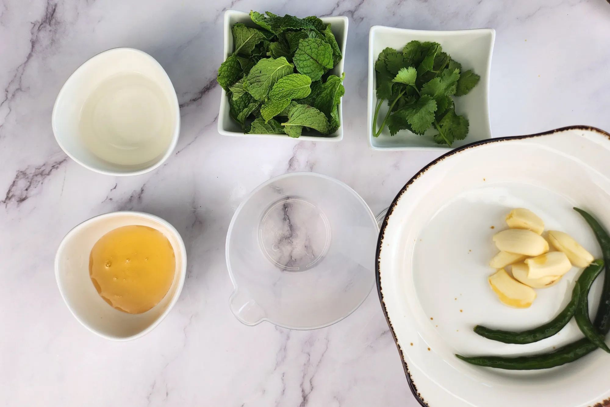 A plate of garlic and bird's eye chilis above a mixing jar surrounded with mint, cilantro, vinegar and honey.