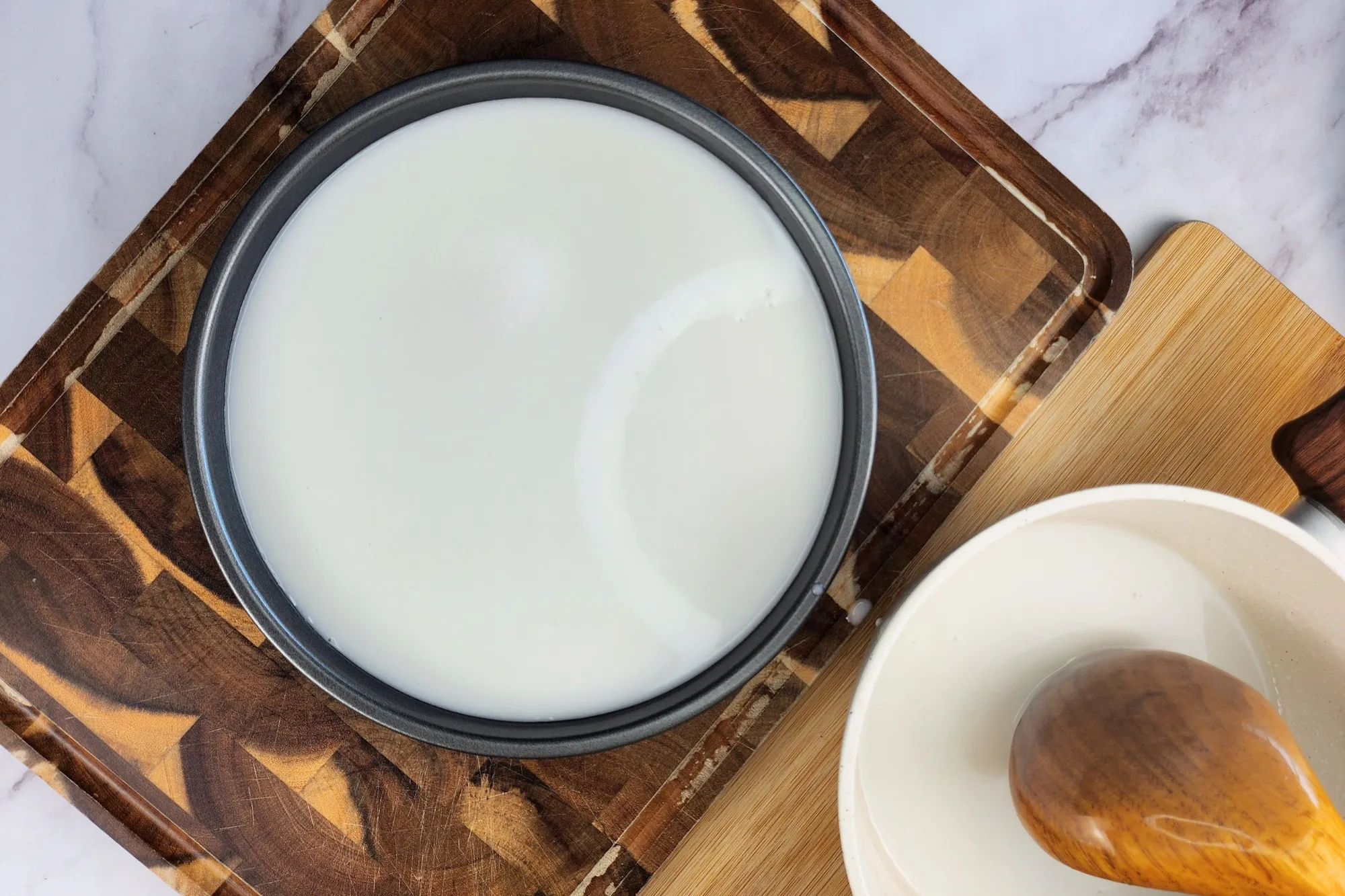 A round tray of white liquid on a chopping board next to a pot and a wooden spoon