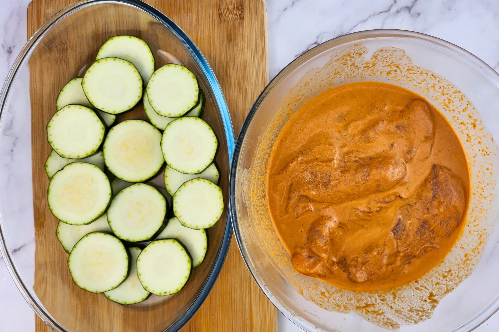Sliced zucchini laid in a baking bowl is next to a marinated chicken in a mixing bowl