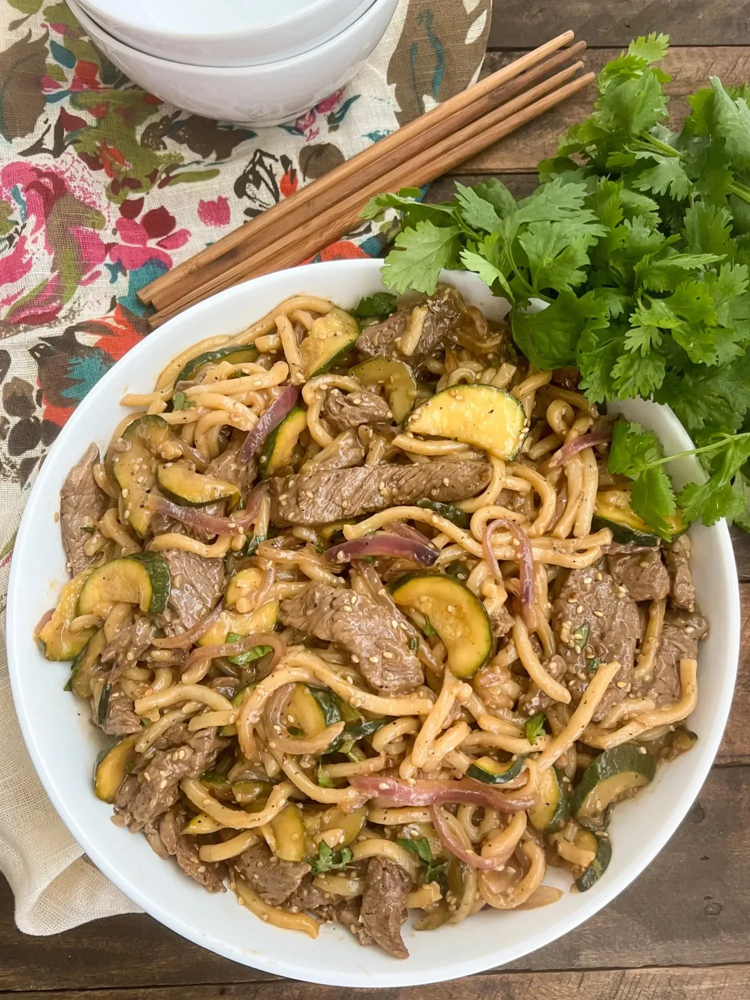 Branded image of Teriyaki Beef Udon Noodles in a white bowl with chopsticks and a cilantro garnish.