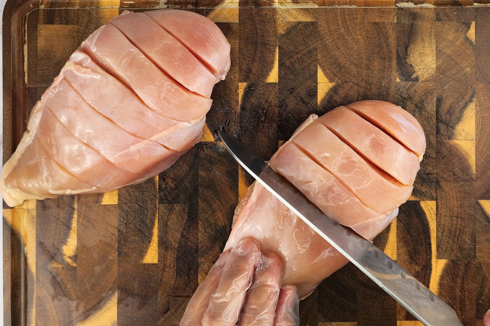 Two cut chicken breasts, a hand holding a knife cut a chicken breast